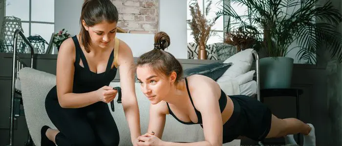 two women indoors. one woman is doing a plank and the second woman is timing her.